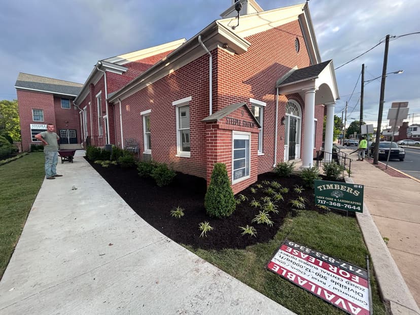 Renovated brick building with landscaping, Timbers sign, and workers mulching nearby.