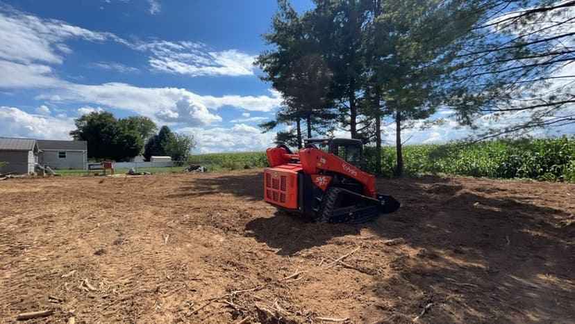 Skid steer loader on cleared land with trees and cornfield under a blue sky.