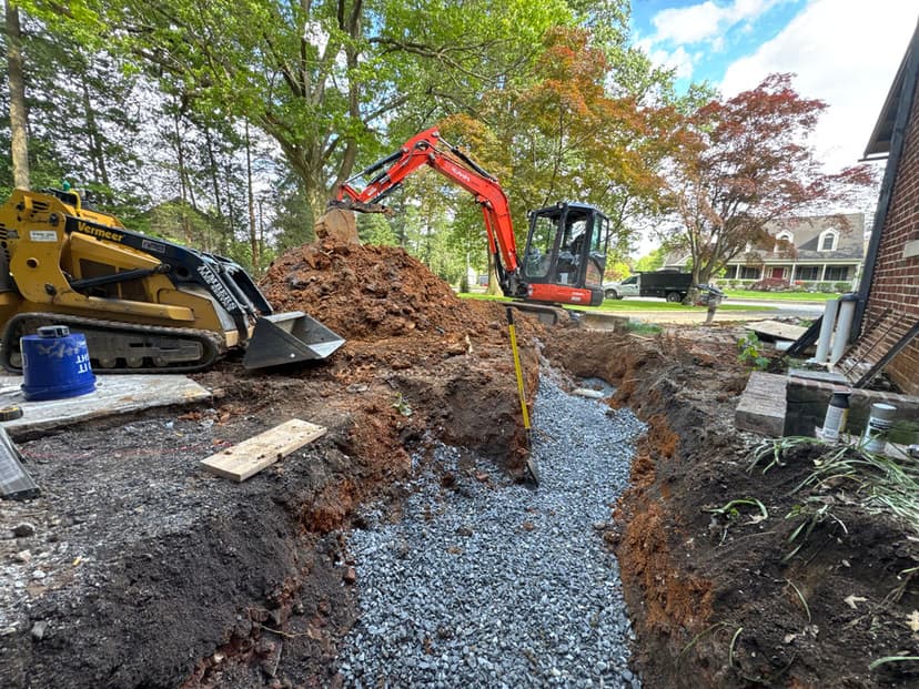 Excavation site with heavy machinery digging a trench and a pile of dirt, surrounded by trees.