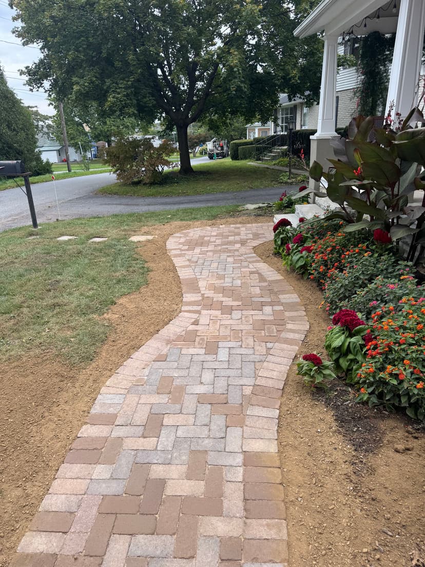 Curved brick pathway leading to a house, surrounded by colorful flower beds and greenery.