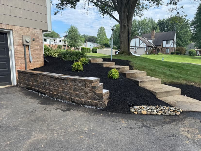 Landscaped garden with stone steps, mulch, and greenery beside a residential driveway.