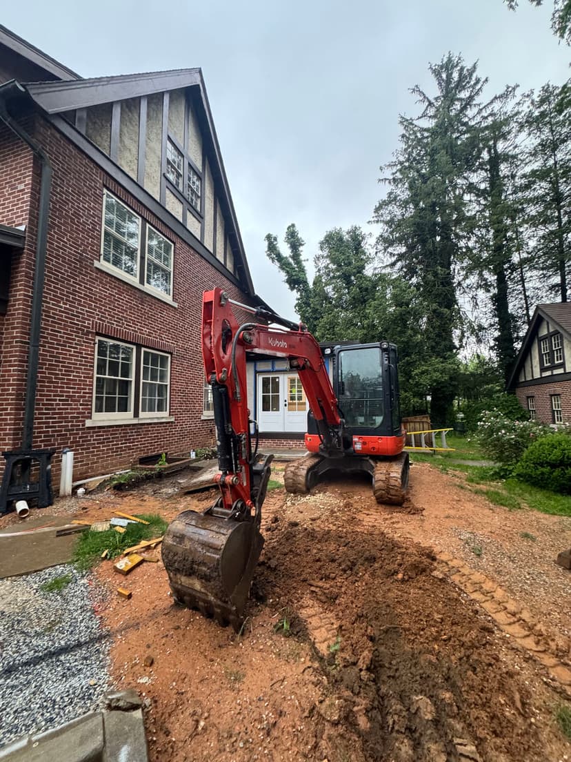 Excavator machine on construction site near brick house with trees in background.