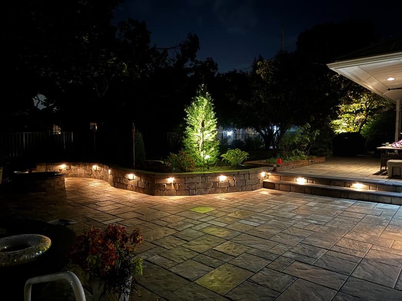 Night view of a patio with stone walls, LED lights, and a lit tree, surrounded by greenery.
