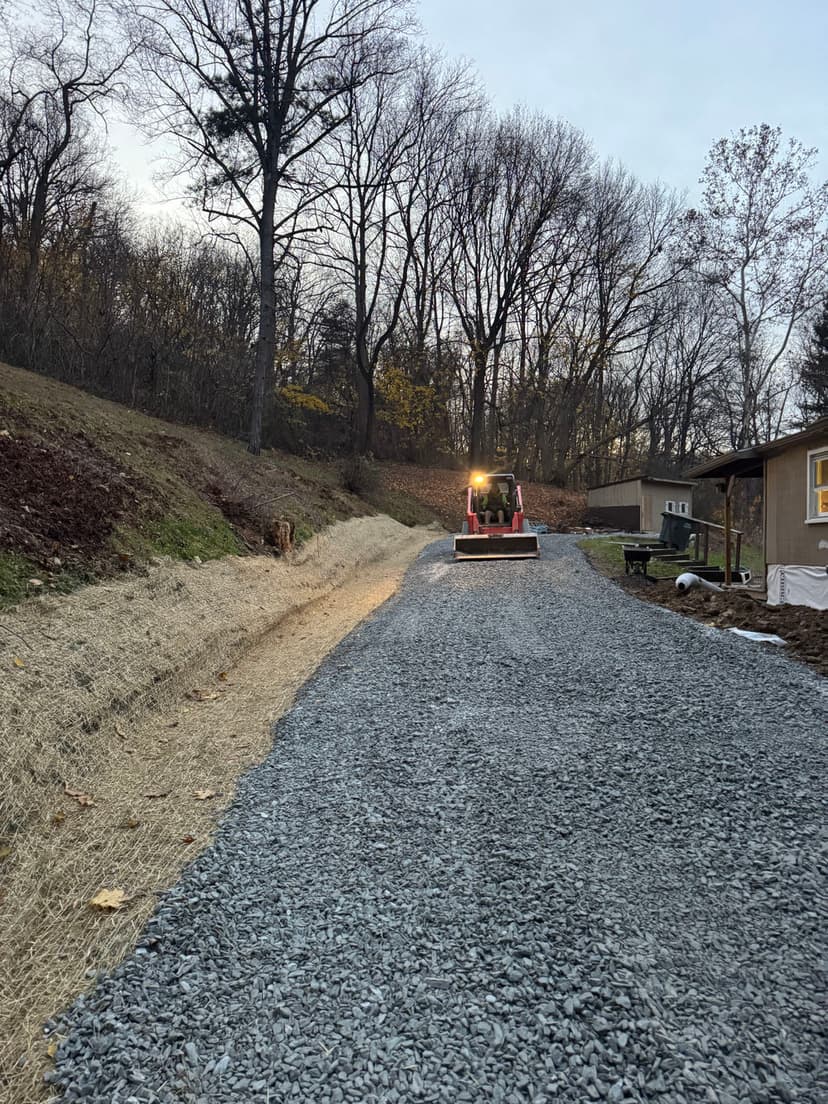 Gravel driveway with tractor working on hillside surrounded by trees at sunset.