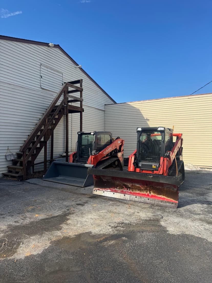 Two orange skid steer loaders with plows parked beside a building under a clear blue sky.