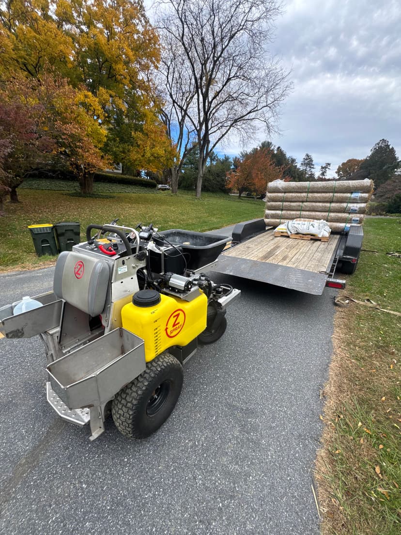 Lawn care equipment and rolled sod on a trailer in a residential yard with autumn scenery.