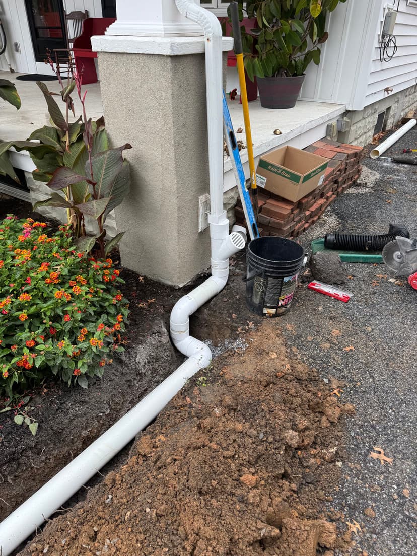 Drainage pipe installation outside a home, with flowers and construction tools nearby.