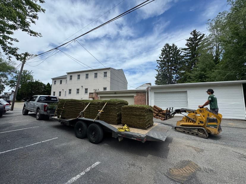 Truck with trailer loaded with sod grass rolls in a residential area. Worker using mini skid steer.