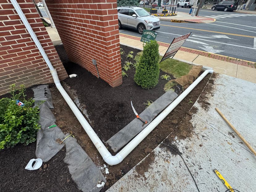 Drainage pipe installation near a brick building with landscaped garden and road view.