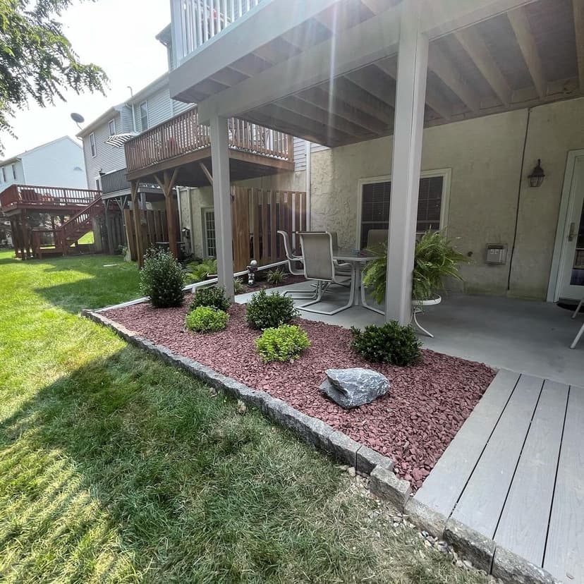 Patio area with seating, landscaped with mulch and shrubs, under a wooden deck.