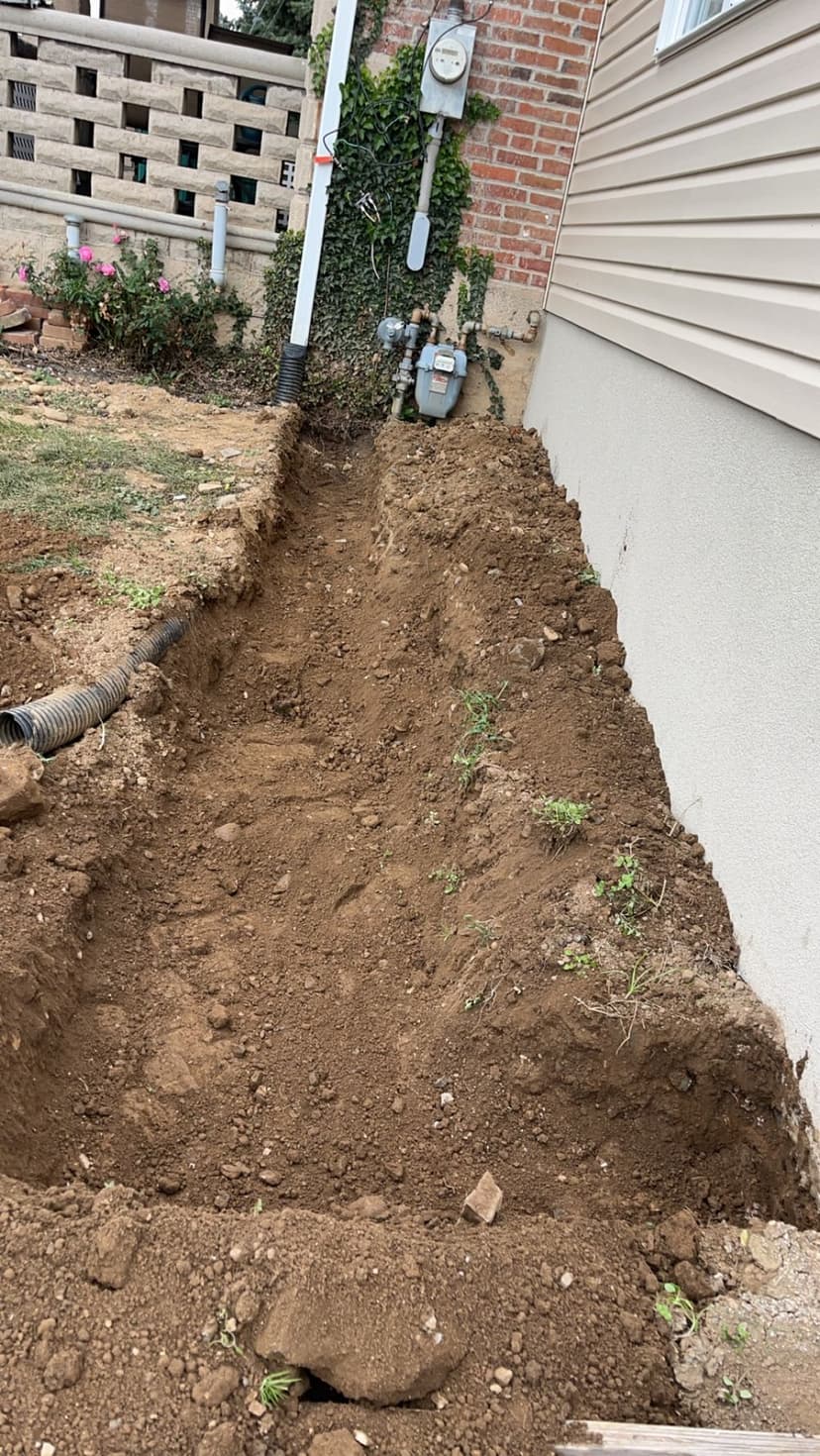 Excavated trench next to house for utility installation, with visible soil and pipes.